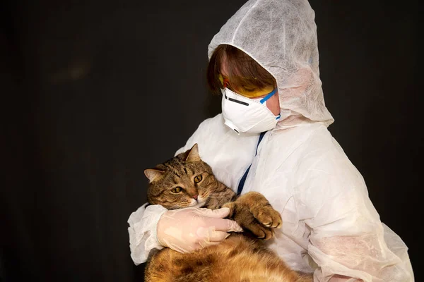 Tıbbi maskeli, koruyucu kıyafetli ve elinde koruyucu sarı gözlüklü bir kız koyu bir arka planda bir kedi tutuyor.               