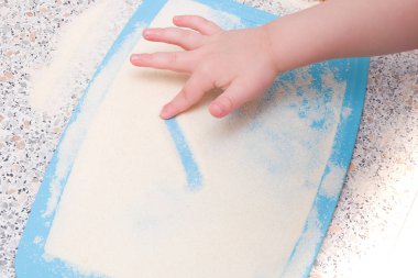 Küçük bir çocuk irmikle bir parmak çizer, mutfak masası, motor becerilerinin gelişimi, bir çocukla ne yapması gerektiği, mavi tahtayı kesmesi.