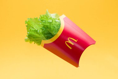 Havada asılı duran ünlü McDonald 's markasının patates kızartması paketinde taze yeşil marul, sarı arka planda uçan nesneler, kırmızı sarı karton kutu, fast food konseptine karşı düzgün beslenme, sağlıklı beslenme.