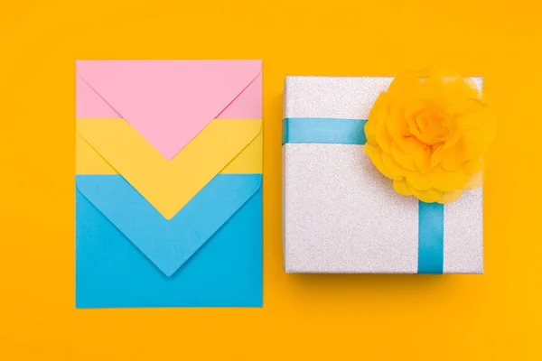 three envelopes of yellow, blue and pink colors nested in each other lie on a yellow background next to a shiny gift box with a yellow flower and blue summer copy space top view