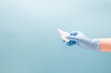 Mavi, tek kullanımlık tıbbi eldivenli bir kadın eli, burun için beyaz plastik bir şişe tutuyor, kapağı yok, mavi arka plan fotokopi alanı, burun damlası, hayır için ilaç.