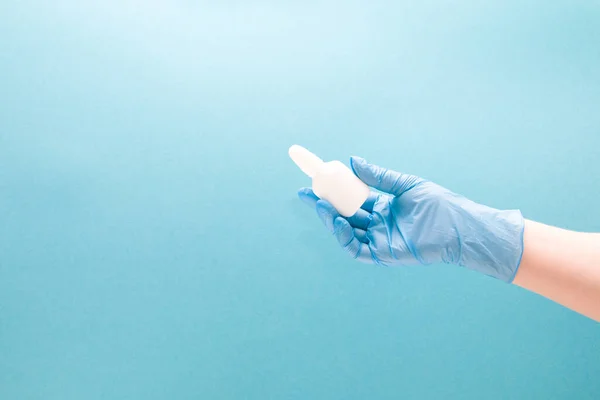 a female hand in a blue disposable medical glove holds a white plastic bottle with drops for the nose, blue background copy space, nasal drops, medicine for the nose