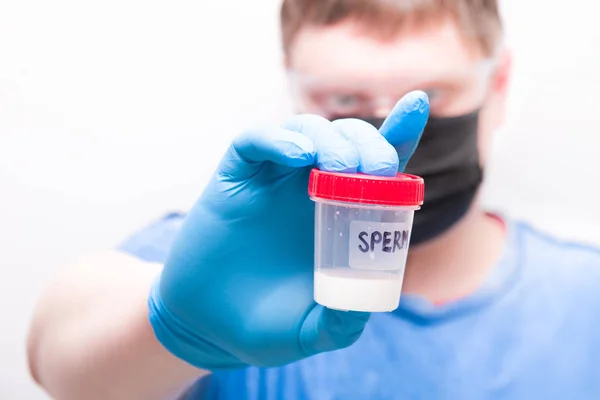 a man in goggles and a medical black mask holds a jar of sprerm in his hand. a hand in a blue disposable glove, a test jar with the inscription \