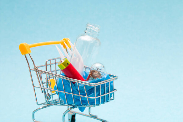 disposable plastic white plug, battery, light bulb and bottle in a shopping trolley on a blue background copy space, we buy trash, zero waste lifestyle concept