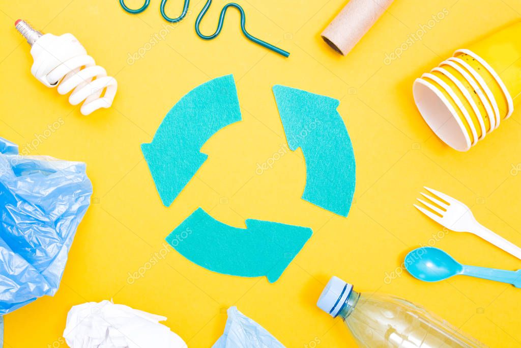 Various trash and a recycling sign in the middle, yellow background, copy space, top view, bottles, disposable tableware, cardboard and paper, eco friendly, plastic bag