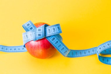 Kırmızı elma yay, kopyalama alanı, sarı arkaplan, diyet ve kilo kaybı kavramına sahip mavi bir ölçüm bandıyla bağlanmış.