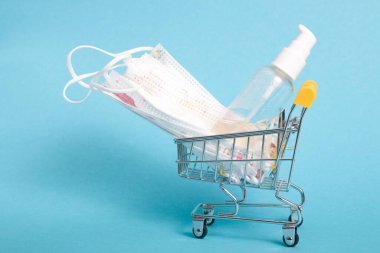 Küçük bir alışveriş arabasında dezenfeksiyon için koruyucu beyaz bebek maskeleri ve jel, mavi arka plan, kopyalama alanı