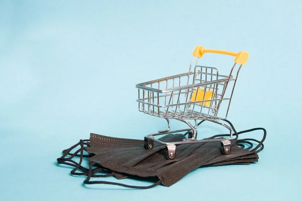 miniature grocery shopping cart stands on black protective medical masks, blue background, copy space, purchase of masks for protection against coronavirus, quarantine concept