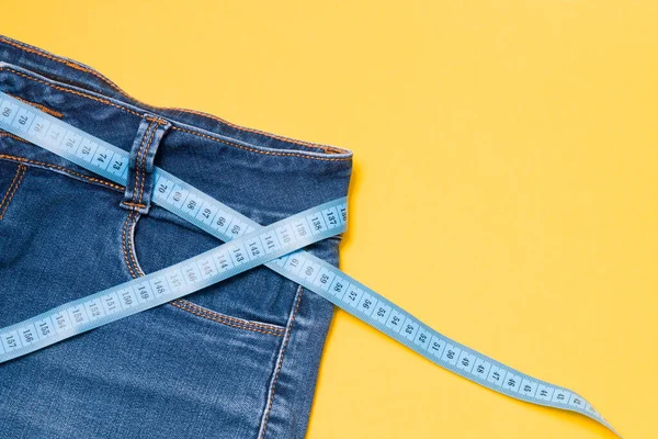 ベルトの代わりにブルーの測定テープをジーンズにねじ込み 黄色の背景にブルーのジーンズ コピースペース 減量と重量管理の概念 測定ボディボリューム — ストック写真