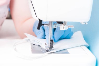 Tek kullanımlık mavi eldivenli bir kadın dikiş makinesine dikiş dikiyor pamuk grisinden yapılmış bir maske, mavi arka plan, dikiş maskesi, dikiş makinesine odaklan.