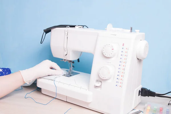 a woman in white disposable medical gloves sews on a sewing machine a protective face mask made of gray cotton fabric, blue background, we sit at home concept, quarantine 2020, shortage of masks