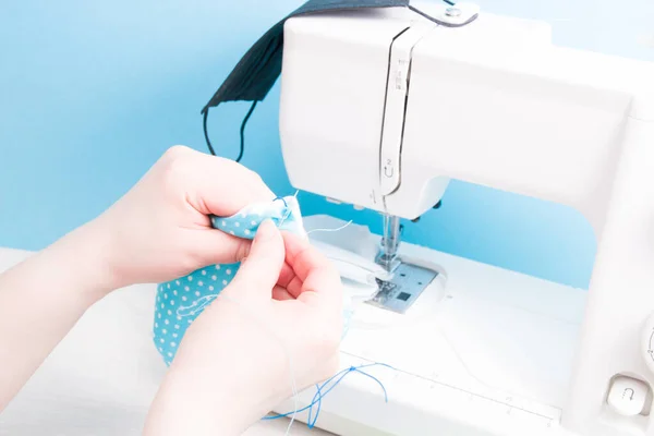 woman sews with a needle a protective face mask made of cotton fabric, hand sewing concept, sewing machine and mask on the background, blue background, virus protection concept, sewing process