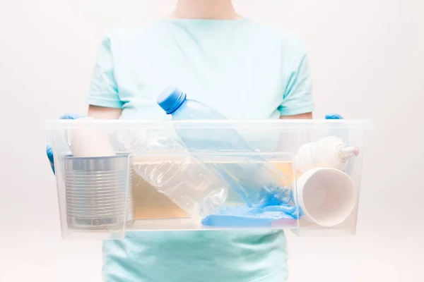 woman in a light blue t-shirt holds a plastic container with waste for recycling, waste-free life style, plastic bottles, a light bulb and a tin can, garbage sorting concept