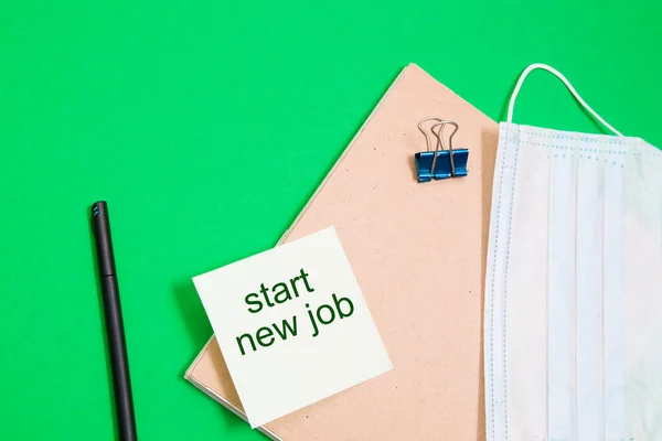 notepad with a recycled cardboard cover and a medical protective mask on a green background, copy space, stationery and green sticker, inscription start a new job, remote work concept