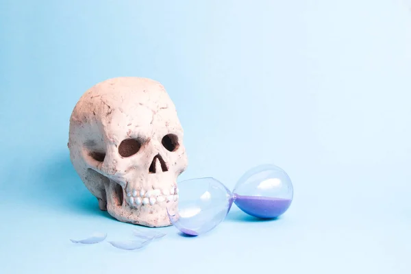 ceramic skull and broken hourglass with purple sand on blue background copy space, death from illness