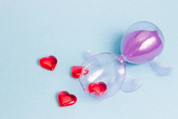 glass broken hourglass with purple sand on and small red glass hearts lie on a blue background, broken heart concept, unhappy love and breakup, pieces of glass, copy space