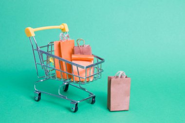 Yeşil arka planda minyatür bir alışveriş arabası, el arabasındaki küçük kağıt poşetler, fotokopi alanı, online alışveriş konsepti, yakınlarda duran bir paket, karantina ve kendini izole etme döneminde mal teslimi.