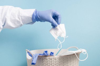Tek kullanımlık mavi eldivenli bir el kullanılmış bir koruyucu maskeyi çöp kutusuna atar, mavi arka plan, fotokopi alanı, beyaz önlüklü bir doktor tramvaylara maske atar.