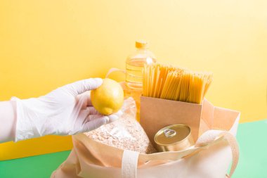 Beyaz plastik bir tek kullanımlık eldiven, yeşil-sarı bir arka plana limon koyar. İçinde bakkaliye olan plastik bir torba, koronavirüs salgınına yardımcı olur. Gönüllü iş konsepti.