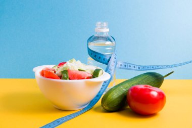 Mavi mezura, bir şişe su, beyaz kasede salata, mavi ve sarı arka planda salatalık ve domates fotokopi alanı, diyet ve kilo kaybı konsepti.