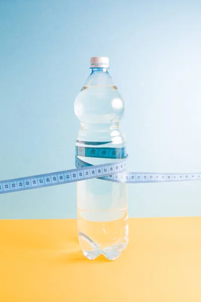 a bottle of water wrapped in a measuring tape stands on a blue-yellow background copy space, healthy life social, support for the body\'s water balance