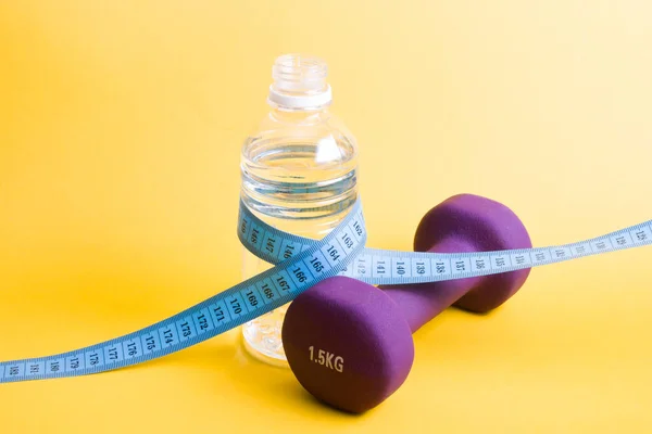 a bottle of water wrapped with a measuring tape and fillet dumbbell on a yellow background copy space, healthy lifestyle social, weight loss and drinking water concept