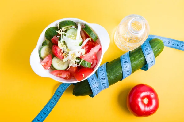 小瓶水 白碗里的沙拉 黄瓜周围的蓝色测量带 蓝色和黄色背景上的西红柿复制空间 饮食和减肥概念 顶视图 — 图库照片