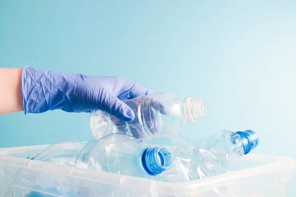 a hand in a blue rubber one-time glove puts an empty plastic bottle of boose water into a container with bottles for plastic recycling, waste sorting concept, blue background, copy space