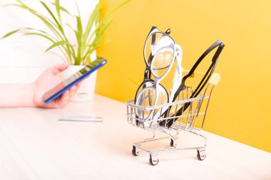 Masadaki küçük alışveriş arabasında görüş düzeltmek için birkaç farklı gözlük, online satın alma gözlüğü, ödeme kartı ve arka planda bulanık bir akıllı telefon olan bir el.