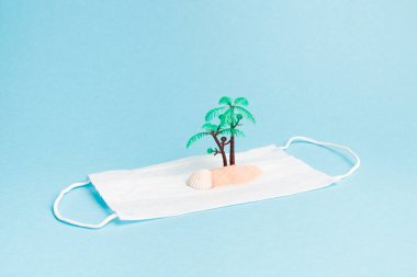 Bir koruyucu yüz maskesi üzerinde avuç içi, kum ve kabuk, mavi arkaplan kopyalama alanı, seyahat ve karantina konsepti, salgın sırasında uçuş yasağı, tatil transferi