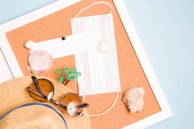 Hasır şapka, tıbbi yüz maskesi, palmiye ağacı, güneş gözlüğü ve mantar panodaki deniz kabukları, metin ışığı için yer, mavi arkaplan, fotokopi yeri, üst görünüm, seyahat ve karantina konsepti