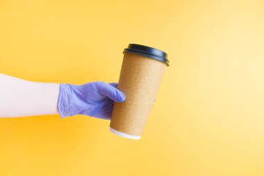 Mavi, tek kullanımlık plastik bir eldiven içinde hazır yemek için parlak, tek kullanımlık bir kahve fincanı, temassız teslimat konsepti, sarı arka plan fotokopi alanı.