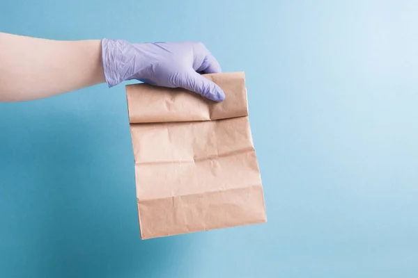 contactless food delivery concept, courier\'s hand in a blue rubber glove holds an order, paper bag on a blue background, copy space