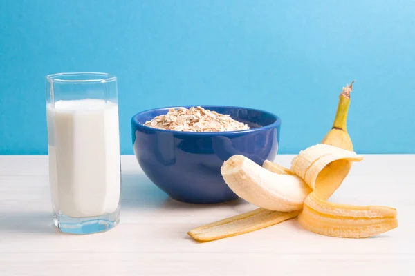 blue bowl with dry oatmeal, staak of milk and banana on a wooden table, blue background, proper nutrition concept, healthy lifestyle, oatmeal with banana for breakfast