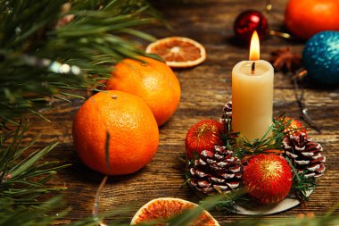 Yanan mumlar, mandalinalar, kozalaklar ve tahta bir masanın üstündeki toplarla Noel süslemeleri.