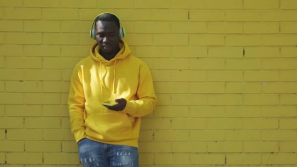 Αφρικανός νεαρός ακούγοντας μουσική ακουμπάει στον κίτρινο τοίχο — Αρχείο Βίντεο