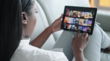 Afro-Amerikalı bir kadın evdeki oturma odasında kanepede uzanmış boş vakit geçirmek için film ya da TV dizisi arıyor. Siyahi kadın internetten film seçiyor. Omuzların üstünden.