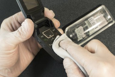 Tek kullanımlık eldivenlerdeki erkek eller siyah arka plandaki eski bir telefondan parmak izlerini silerler. Eldivenli eller eski telefonu dezenfekte eder. Arka planda bakteriyel bir maddeyle dolu bir şişe var.