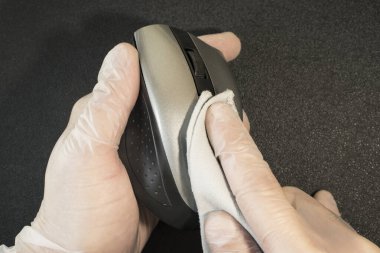 Tek kullanımlık eldivenlerdeki erkek eller siyah arka plandaki kablosuz bir bilgisayar faresinden parmak izlerini siliyor. Eldivenli eller bir bilgisayar faresini dezenfekte eder