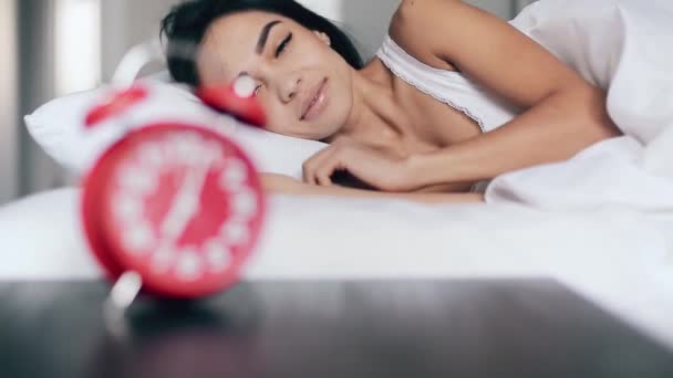 Красивая счастливая девушка просыпается в постели, выключает будильник и встает с кровати — стоковое видео
