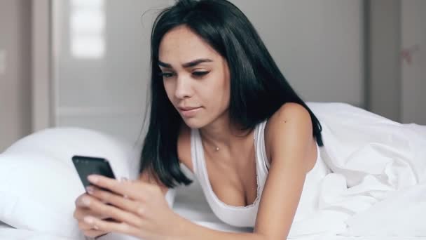 Menina bonita na cama lê mensagens no smartphone e sorrisos — Vídeo de Stock