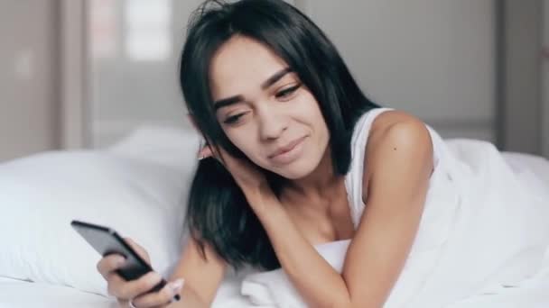 Chica bonita en la cama lee mensajes en el teléfono inteligente y sonríe — Vídeo de stock