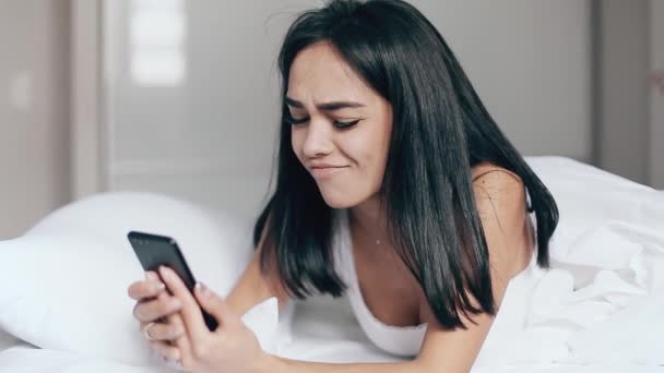 Chica bonita en la cama lee mensajes en el teléfono inteligente y sonríe — Vídeo de stock