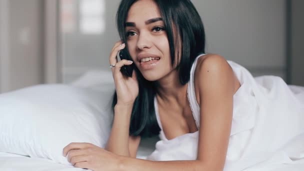 Красива молода брюнетка, посміхаючись брехати, розмовляє мобільний телефон, розслабляється в спальні — стокове відео