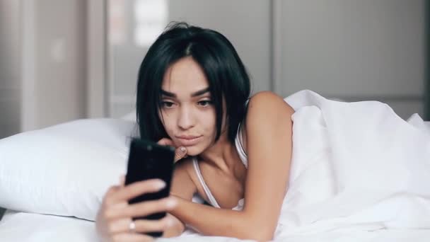 Bonita chica joven en una cama toma selfies usando un teléfono inteligente — Vídeos de Stock
