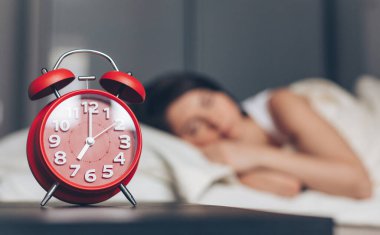 Komodinin üzerinde kırmızı alarm saati ve arka planda uyuyan kadın..
