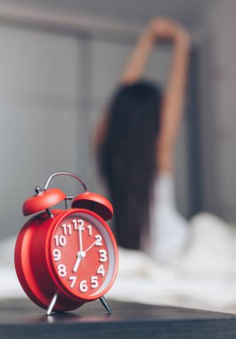 Arka planda mutlu bir kadının yatakta esnediği yakın plan çalar saat.