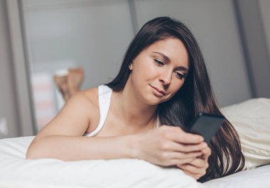 Yatağında yatarken akıllı telefon kullanan mutlu güzel kadın. Gülümseyen esmer kız elinde cep telefonu tutuyor.