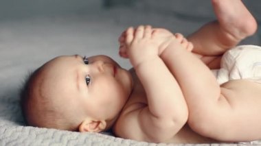 Sevimli gülümseyen beyaz, 6 aylık erkek bebek ayaklarını tutuyor ve yatakta yatarken kameraya bakıyor.