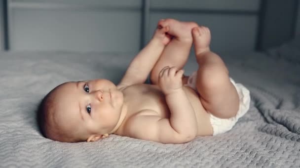 Sevimli gülümseyen beyaz, 6 aylık erkek bebek ayaklarını tutuyor ve yatakta yatarken kameraya bakıyor. — Stok video
